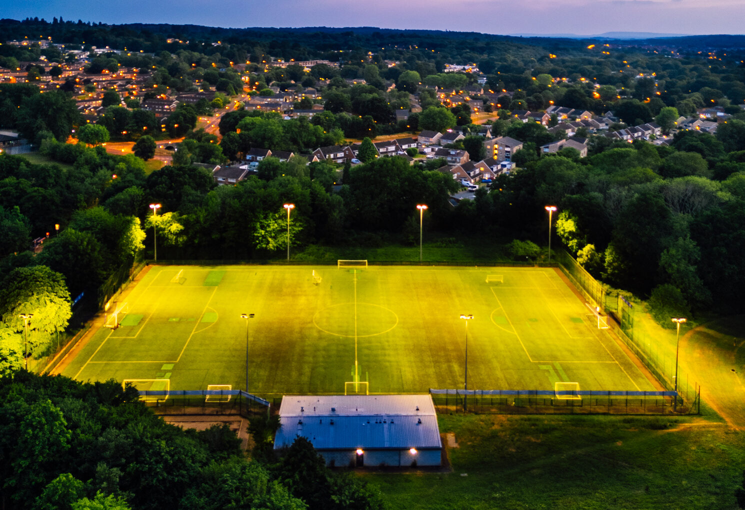 Aerial view, captured by drone, depicting a football pitch illuminated at night with no people. The pitch is surrounded by the streets and houses of a town in southeast England.